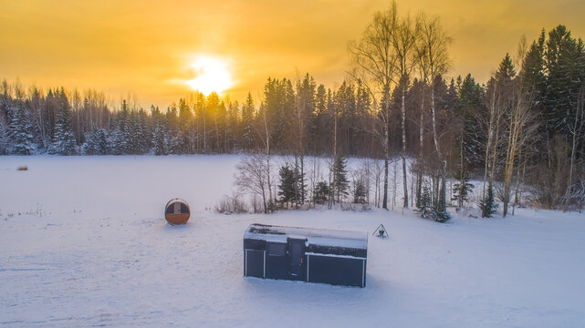 Aerial view of a small dark cabin and a barrel sauna in a snow-covered landscape with a forest backdrop during a golden sunset in Latvia.