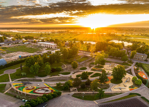 Aerial view of the Jelgava park area with a colorful playground, lush green trees, and the Lielupe river in the background under a golden sunset sky in Jelgava, Latvia.