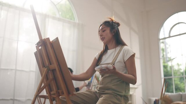 Creative young Asian women friends painting on canvas together in bright home studio. Relaxed female artists enjoying peaceful indoor leisure time, expressing imagination and passion for art.