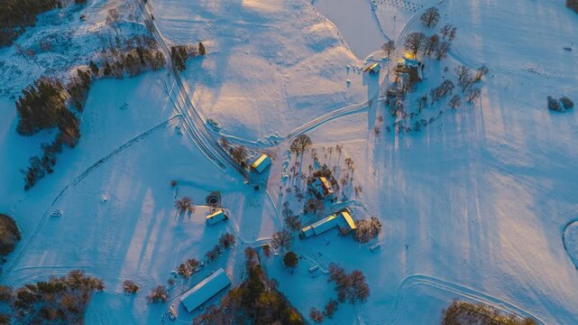 Aerial view of a snow-covered farmstead with outbuildings and long shadows cast across frozen fields during the golden hour in Latvia.