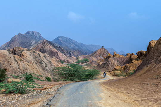 View of a winding road through the arid, rocky Sulaiman Mountains with a person on a motorcycle, sparse shrubs, and clear blue sky in De-excluded Area Dera Ghazi Khan, Punjab, Pakistan.