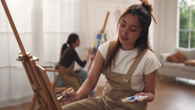 Creative young Asian women friends painting on canvas together in bright home studio. Relaxed female artists enjoying peaceful indoor leisure time, expressing imagination and passion for art.