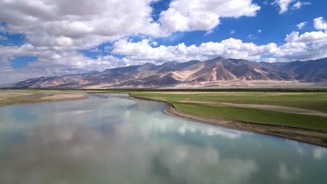 Aerial view of the Indus River flowing through the green plains of Dungti with the Himalayan mountains in the background under a cloudy blue sky in Dungti, Ladakh, India.