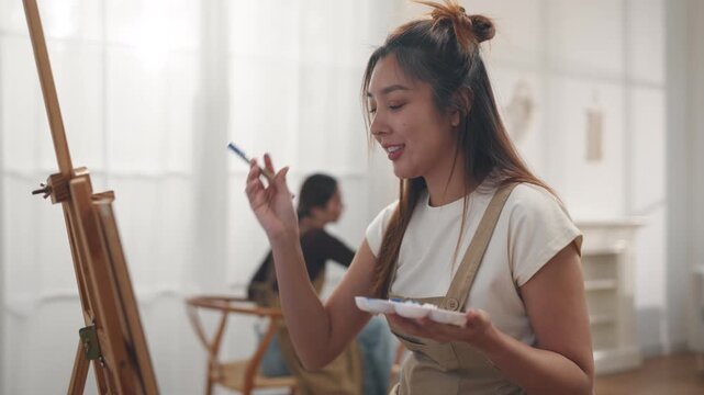 Creative young Asian women friends painting on canvas together in bright home studio. Relaxed female artists enjoying peaceful indoor leisure time, expressing imagination and passion for art.