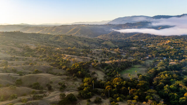 Aerial view of rolling hills, lush landscape with trees, morning mist over mountains, and golden sunlight in San Jose de Las Matas, Santiago Province, Dominican Republic.