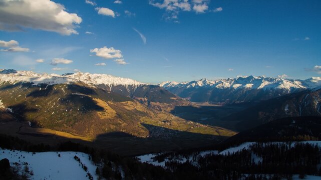 Aerial view of the Watles mountain area with snow-capped peaks, deep valleys, and pine forests under a clear blue sky in Mals, Trentino-Alto Adige, Italy.