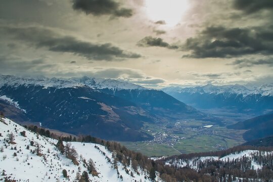 Aerial view of Watles mountain peak overlooking the snow-covered valley and the town of Mals, with dramatic clouds and sunlight in Mals, Trentino-Alto Adige, Italy.