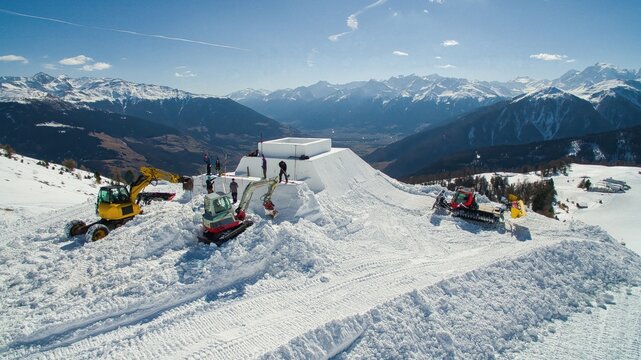 Aerial view of Watles with excavators and snow groomers shaping a large snow structure on a sunny day in the mountains of Mals, Trentino-Alto Adige, Italy.