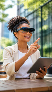 Young woman wearing AR smart glasses and using tablet outdoors, pointing at virtual interface.