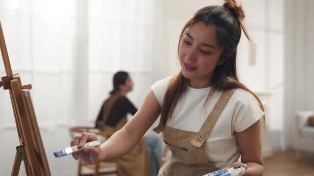 Creative young Asian women friends painting on canvas together in bright home studio. Relaxed female artists enjoying peaceful indoor leisure time, expressing imagination and passion for art.