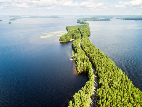 Aerial view of Pulkkilanharju ridge, a scenic esker formation with a road winding through dense pine forests, surrounded by deep blue waters of Lake Paijanne in Asikkala, Paijat-Hame, Finland.