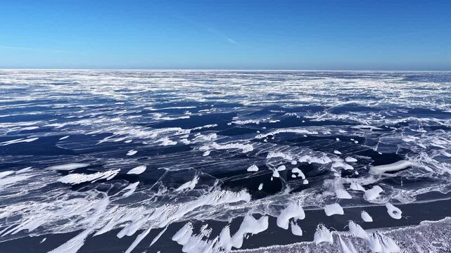 Aerial view of frozen cracked ice patterns on a vast lake surface under a clear blue sky in Latvia.
