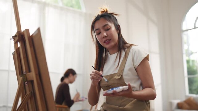 Creative young Asian women friends painting on canvas together in bright home studio. Relaxed female artists enjoying peaceful indoor leisure time, expressing imagination and passion for art.