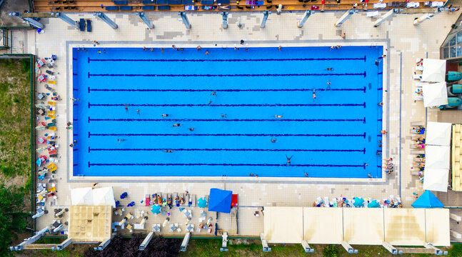 Aerial view of a rectangular swimming pool with people swimming in the lanes, surrounded by lounge chairs, umbrellas, and a tiled deck area in Novi Sad, Vojvodina, Serbia.