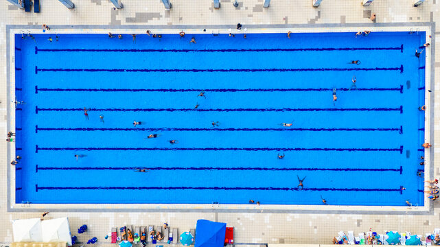 Aerial view of a large rectangular swimming pool with multiple lanes and people swimming, surrounded by a tiled deck with lounge chairs and umbrellas in Novi Sad, Vojvodina, Serbia.