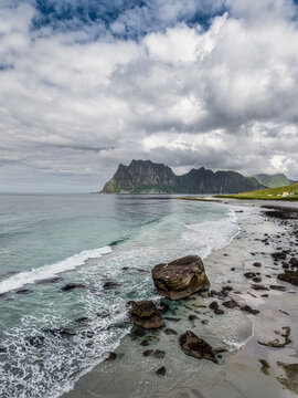 Aerial view of Uttakleiv Beach with large rocks on the sandy shore, turquoise sea waves, and dramatic mountains under a cloudy sky in Leknes, Nordland, Norway.