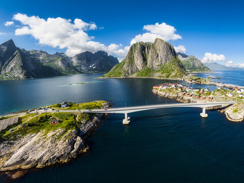Aerial view of the iconic Lofoten bridge connecting islands with red houses, rocky mountains, and deep blue water in Moskenes, Nordland, Norway.