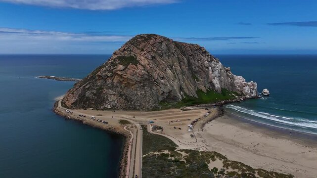 Aerial view of Morro Rock rising from the Pacific Ocean with a sandy beach and coastal road in Morro Bay, California, United States.