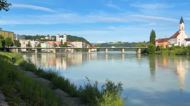 LTR pan across the Passau, Germany bank of the Inn River to the opposite shore, with a view of an auto bridge and a church reflected in the water, on a sunny summer day.
