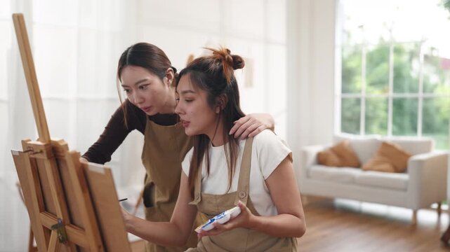 Creative young Asian women friends painting on canvas together in bright home studio. Relaxed female artists enjoying peaceful indoor leisure time, expressing imagination and passion for art.