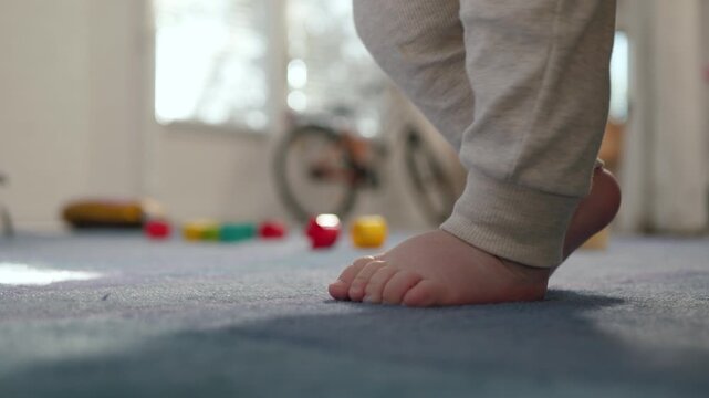 Low-angle close-up showing a toddler's bare feet and lower legs on a plush carpet in a sunlit playroom. Baby begins to walk joyfully, tiny feet taking first happy steps