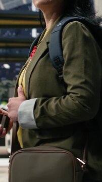 Vertical composition: Upbeat woman holds a mobile phone while moving through a bus station waiting area; the passenger checks route details on the phone as she walks to the coach departure platform.