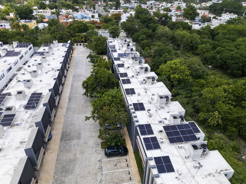 Aerial view of private solar panels installed on residential rooftops in a modern neighborhood with lush green trees and a paved street in Merida, Yucatan, Mexico.