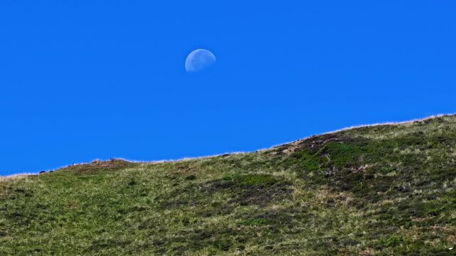 Aerial view of the grassy mountain slope at Col d'Aubisque under a clear blue sky with the moon visible in Beost, Nouvelle-Aquitaine, France.