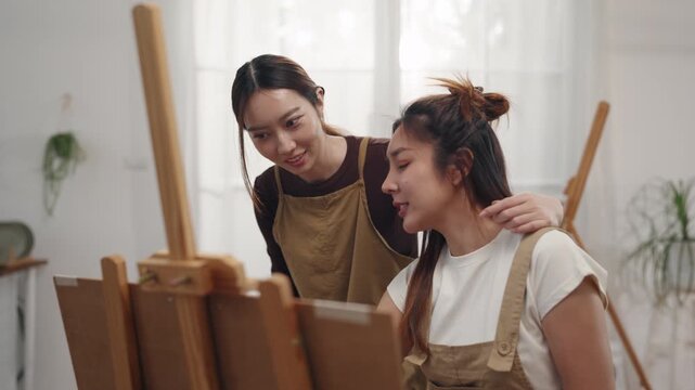 Creative young Asian women friends painting on canvas together in bright home studio. Relaxed female artists enjoying peaceful indoor leisure time, expressing imagination and passion for art.