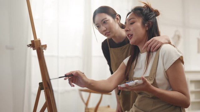 Creative young Asian women friends painting on canvas together in bright home studio. Relaxed female artists enjoying peaceful indoor leisure time, expressing imagination and passion for art.