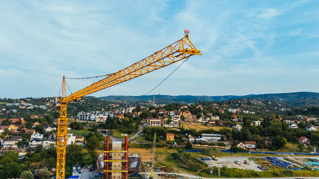 Aerial view of a large yellow tower crane at a construction site with residential buildings and green hills in the background in Novi Sad, Serbia.