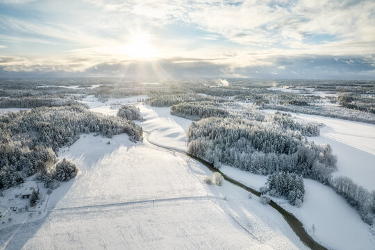 Aerial view of a snow-covered winter landscape with dense pine forests and a winding river under a bright sunburst in Lieto, Southwest Finland, Finland.