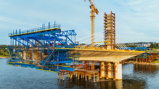 Aerial view of a bridge under construction over a river, featuring large blue steel scaffolding, a tall yellow crane, and concrete supports in Novi Sad, Serbia.