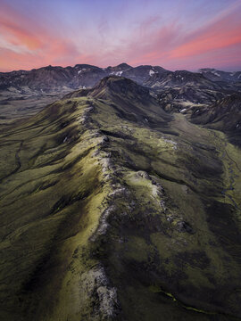 Aerial view of a dramatic mountain ridge covered in vibrant green moss under a vivid pink and orange sunset sky in Iceland.