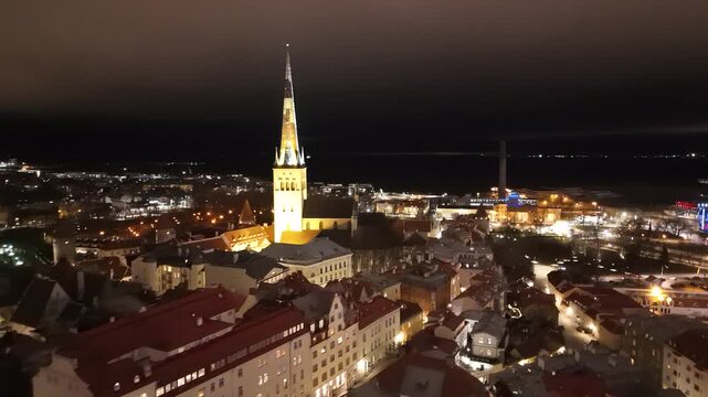 Aerial view of the illuminated St. Olaf's Church spire rising above the historic rooftops of the old town at night in Tallinn, Harju County, Estonia.