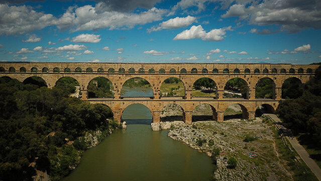 Aerial view of the ancient Roman aqueduct bridge Pont du Gard spanning the Gardon River, surrounded by lush greenery and rocky banks in Vers-Pont-du-Gard, Occitanie, France.