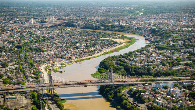 Aerial view of Rio Ozama winding through the dense urban landscape of Santo Domingo, featuring cable-stayed bridges, lush riverbanks, and residential areas. Santo Domingo, DN, Dominican Republic.