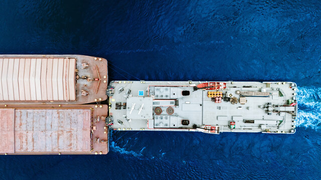 Aerial view of a tugboat pushing a large cargo barge through deep blue sea waters in Greece.