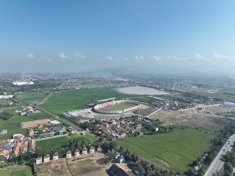Aerial view of Krakatau Steel Stadium surrounded by green rice fields, residential areas, and distant mountains under a clear blue sky in Cilegon, Banten, Indonesia.