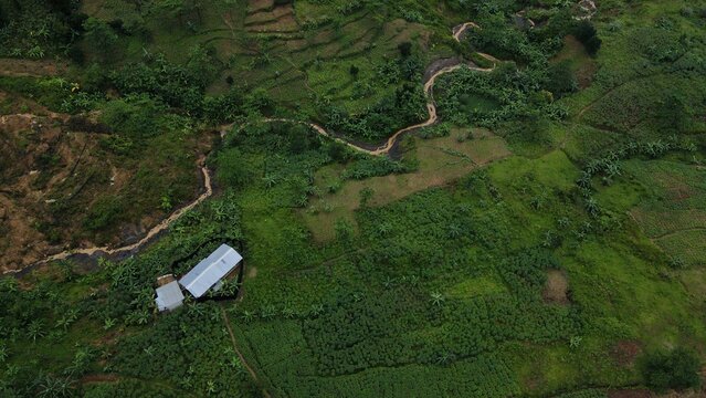 Aerial view of lush green terraced fields and a small stream winding through the rural landscape near a building with a metal roof in Babakan Madang, West Java, Indonesia.