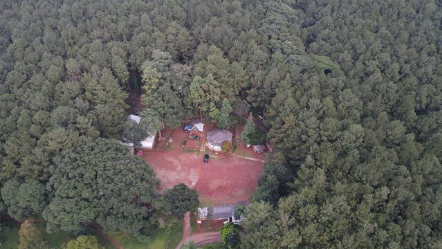 Aerial view of a clearing in a dense pine forest featuring small buildings and a red dirt ground in Babakan Madang, West Java, Indonesia.