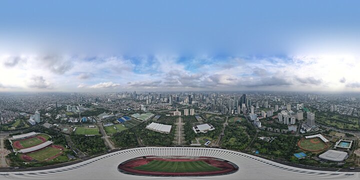 Aerial view of Gelora Bung Karno Stadium surrounded by lush greenery and the sprawling cityscape under a cloudy sky in Kecamatan Tanah Abang, Daerah Khusus Ibukota, Indonesia.
