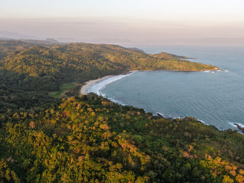 Aerial view of Pantai Legon Pari with lush green hills, sandy beach, and waves crashing on the shore in Kecamatan Bayah, Banten, Indonesia.