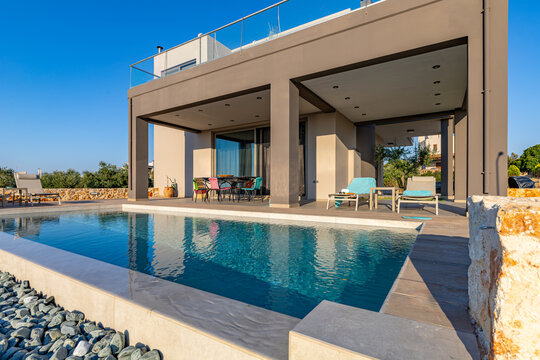 View of a modern luxury villa with a private swimming pool, outdoor dining area, and stone wall in Chania, Greece.