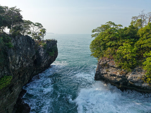 Aerial view of Karang Bokor Sawarna with crashing waves hitting the rocky cliffs and lush green trees in Bayah, Banten, Indonesia.