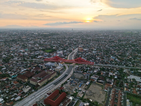 Aerial view of the red Joglo Railway Bridge spanning over the urban landscape with residential rooftops and roads under a soft sunset sky in Kecamatan Banjarsari, Jawa Tengah, Indonesia.