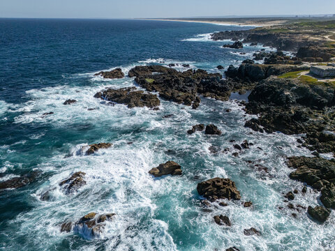 Aerial view of the rugged coastline with dark volcanic rocks and crashing white waves in the Atlantic Ocean near Sines, Setubal, Portugal.