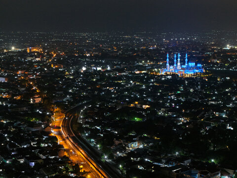 Aerial view of Jembatan Kereta Api Joglo illuminated at night, with the surrounding city lights and a brightly lit mosque in Kecamatan Banjarsari, Jawa Tengah, Indonesia.