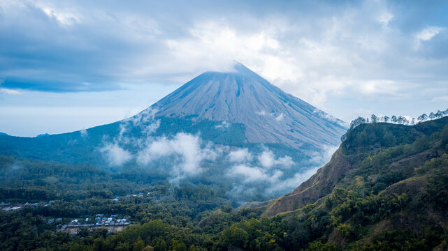 Aerial view of Mount Inerie volcano rising above lush green forested slopes and a small village under a cloudy sky in Inerie, East Nusa Tenggara, Indonesia.
