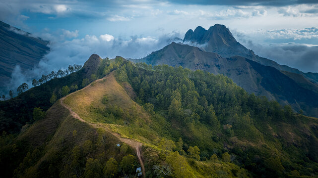 Aerial view of the lush green mountain slopes and a winding dirt path leading towards the dramatic peaks of Inerie volcano surrounded by clouds in Bajawa, Nusa Tenggara Timur, Indonesia.